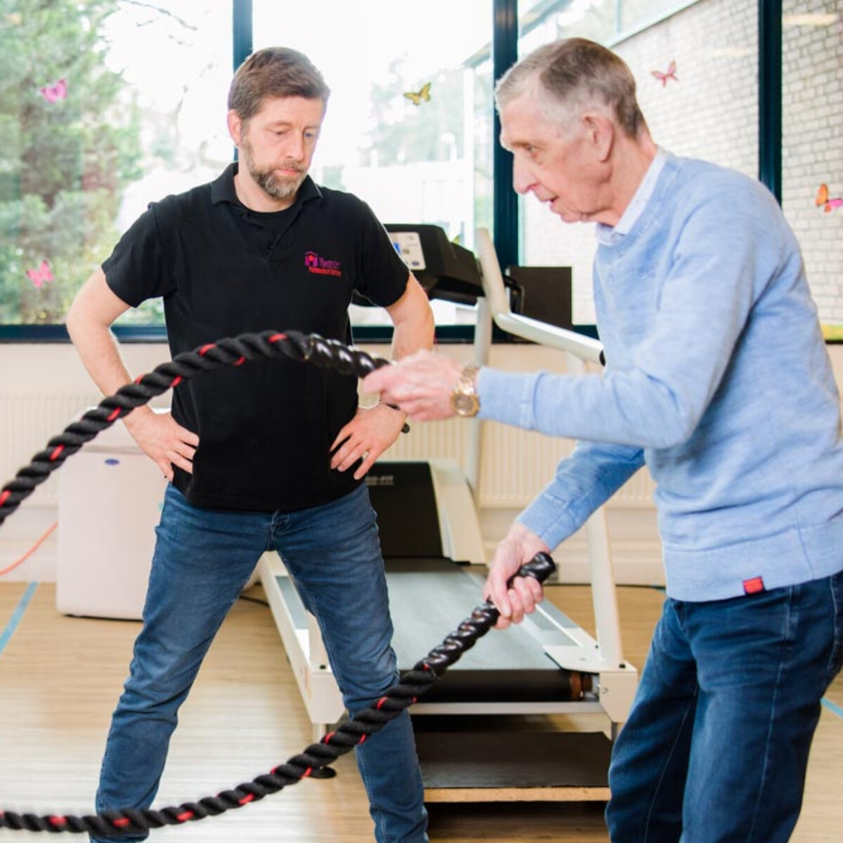 Oude man traint met battle rope onder begeleiding van een instructeur in een sportschool met loopband op de achtergrond.