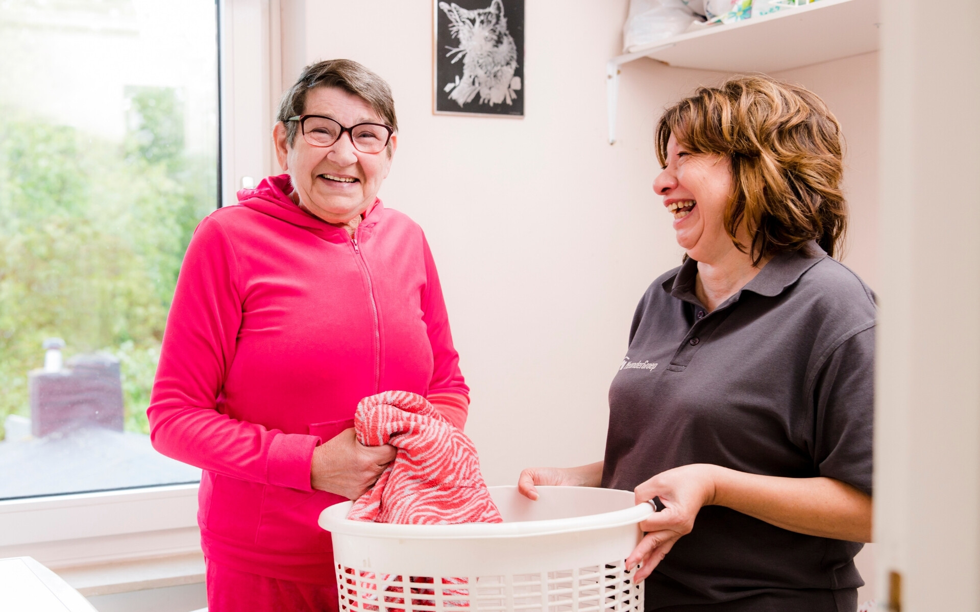 Two women share a moment of laughter in a cozy kitchen, with one holding a red and white striped towel and the other holding a white basket.