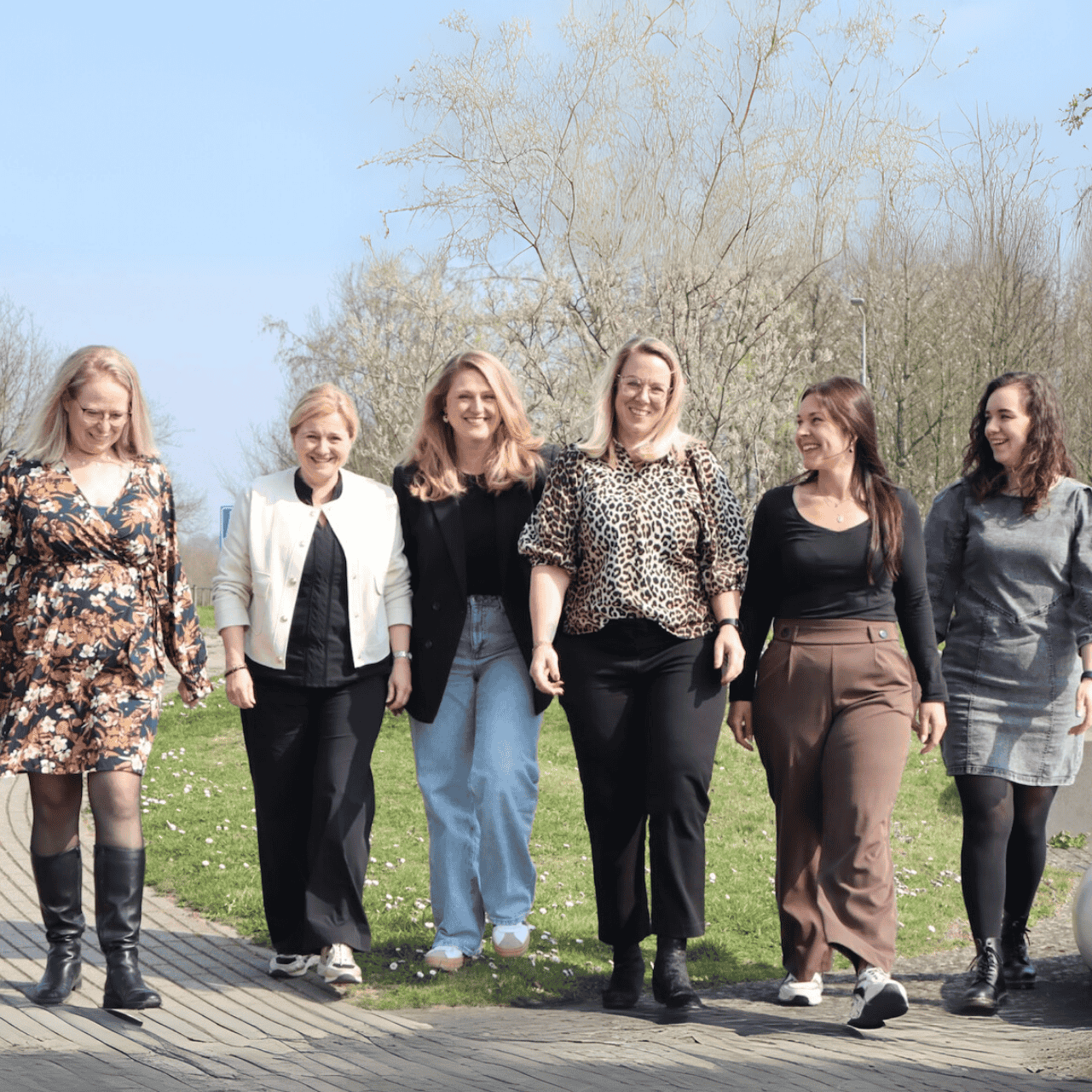 Six women in diverse attire, including jeans, dresses, and boots, walk together on a brick path in a park.