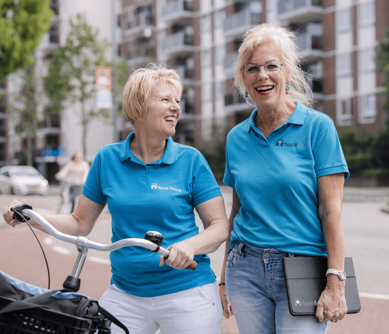 Twee lachende oudere vrouwen in blauwe polo’s met logo’s, één met een fiets, de ander met een tablet, staand in een stedelijke omgeving.