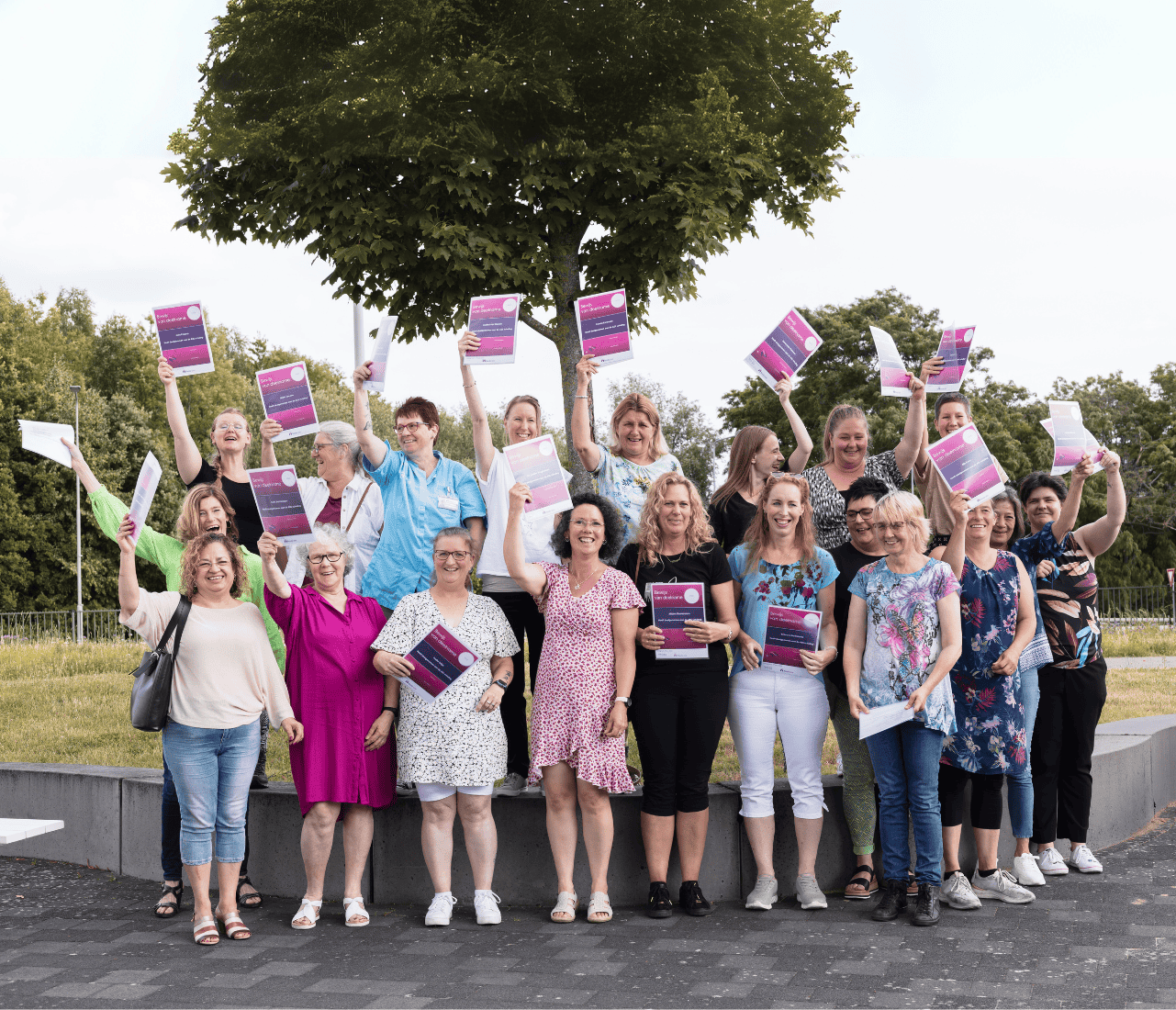 A group of diverse individuals holds purple signs with white text, gathered in a park with a tree and concrete wall in the background.
