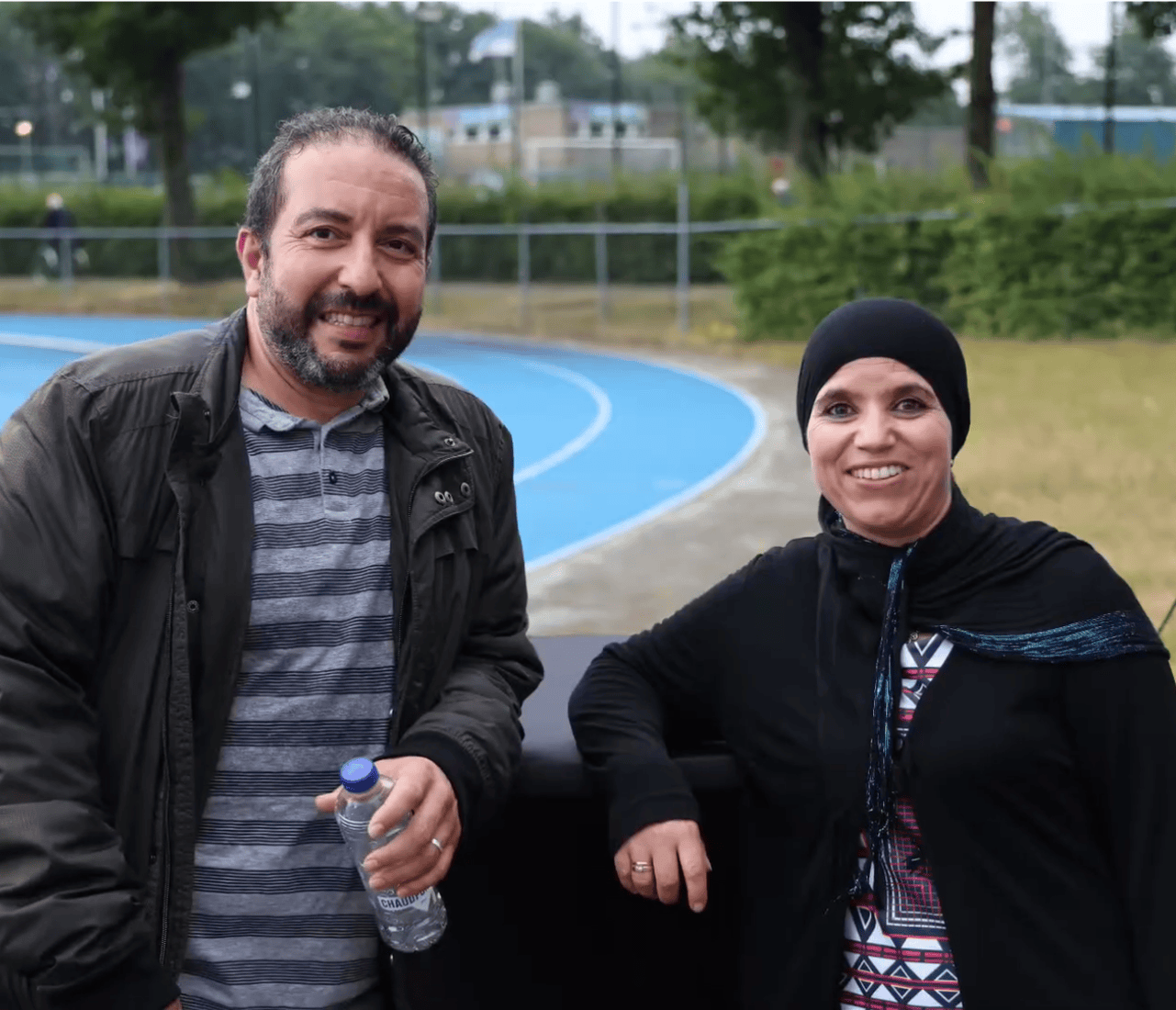 Two individuals, one in a black jacket and gray striped shirt, the other in a black hijab and patterned top, stand near a blue track holding a water bottle.