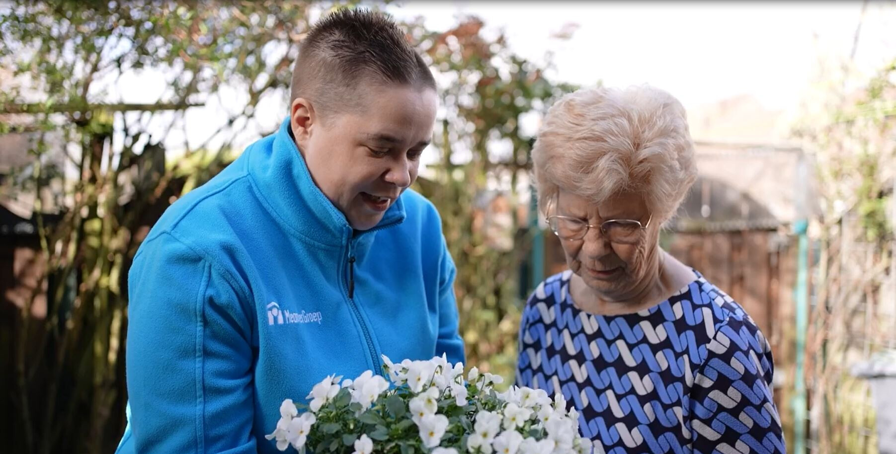 Two individuals interact in a lush garden, with one holding a bunch of white flowers and the other wearing a blue and white patterned shirt.