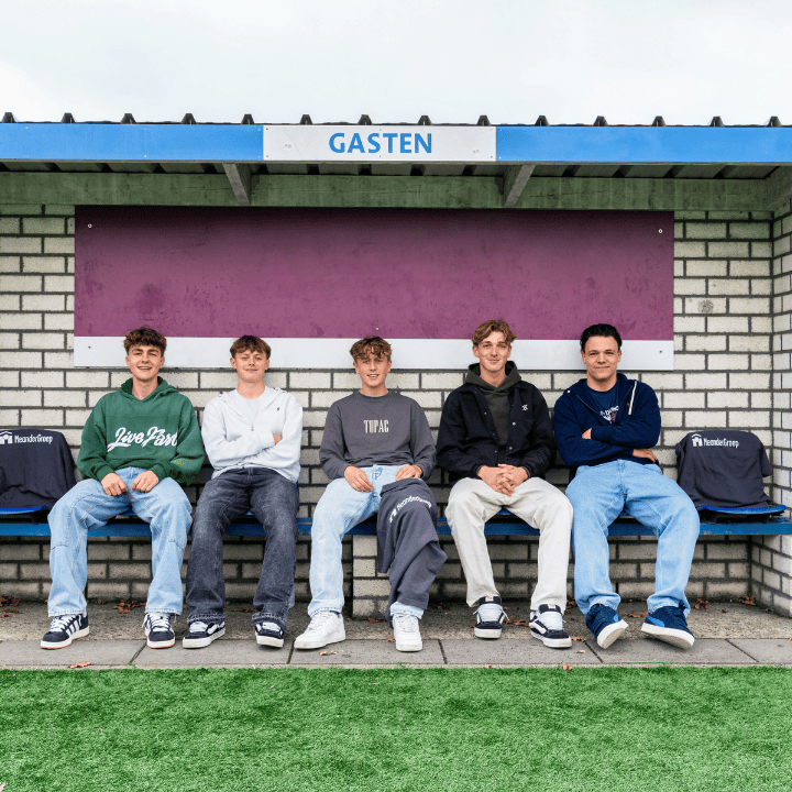 Vijf jonge mannen zittend op een bankje onder een afdakje met bordje "GASTEN", grasveld op de voorgrond.