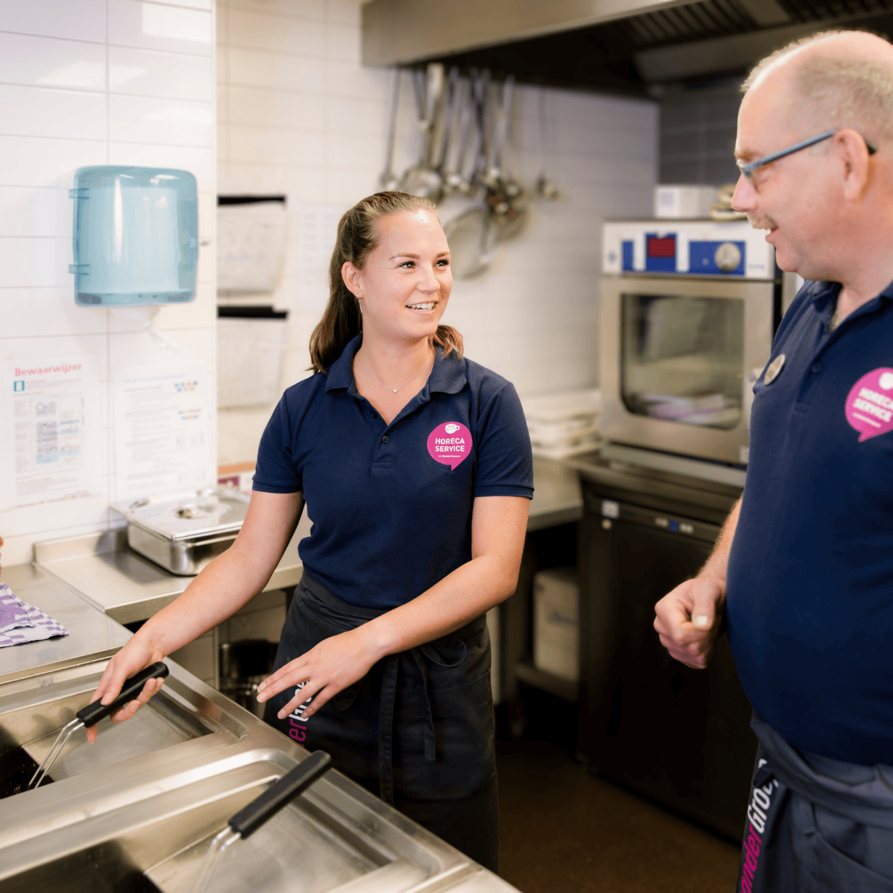 Two individuals in blue polo shirts, one with a pink badge and the other with a pink "Support" badge, work in a kitchen with white tiled walls, a sink, and a stove.