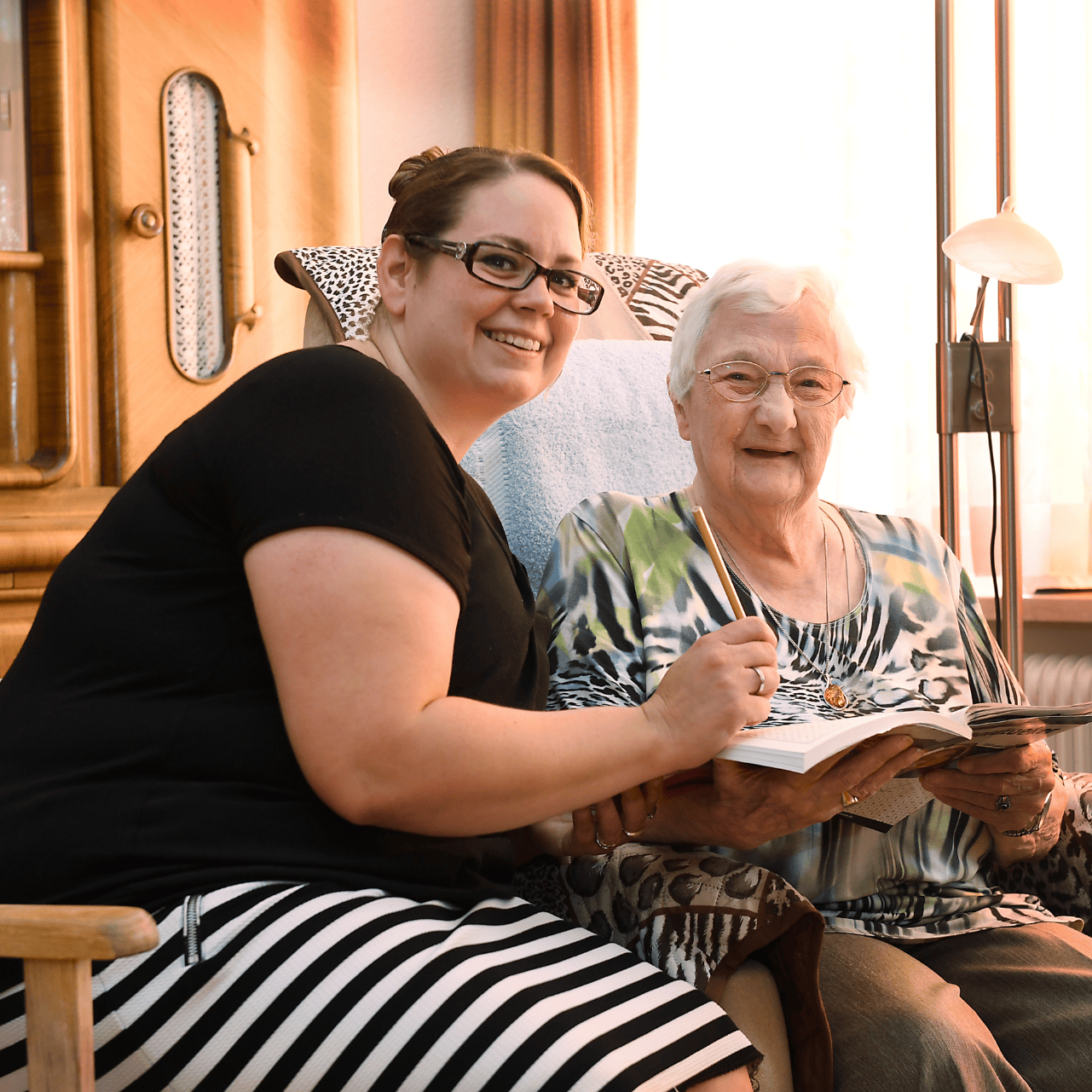 A young woman in a black top and striped skirt sits with a smiling elderly woman in a blue and green blouse, holding a book and pen in a cozy room.